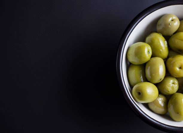 Marinated whole green olives in white metal bowl with black edge isolated on black background
