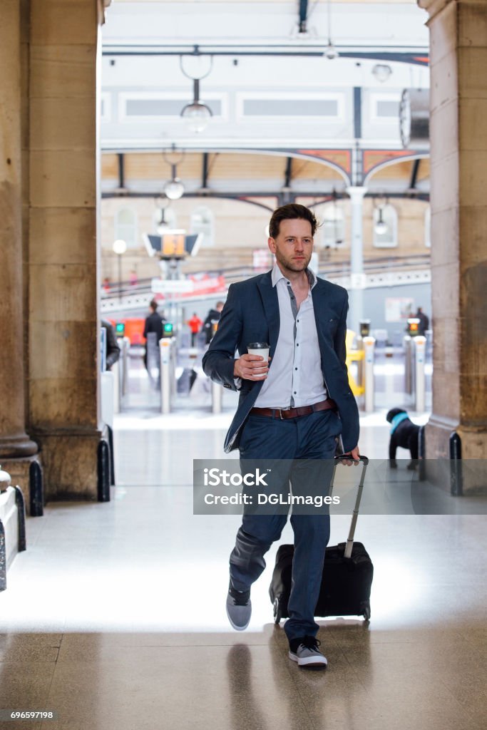 Rushing To Work Businessman is in a hurry as he leaves the train station to commute the work. He is carrying a coffee and pulling a suitcase. Town Stock Photo