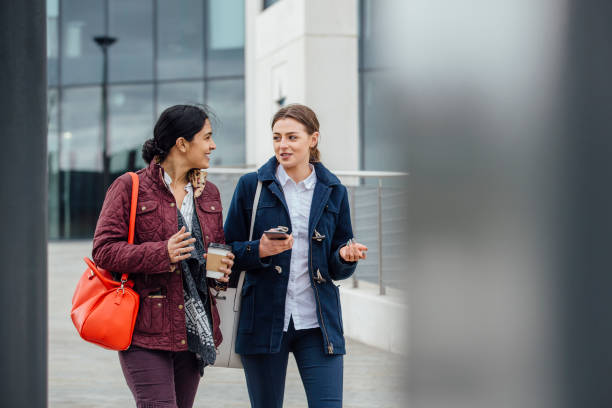 Women Going To Work Businesswomen are walking to work together through the city. They are talking and one is holding a coffee while the other is holding a smartphone. uk street stock pictures, royalty-free photos & images