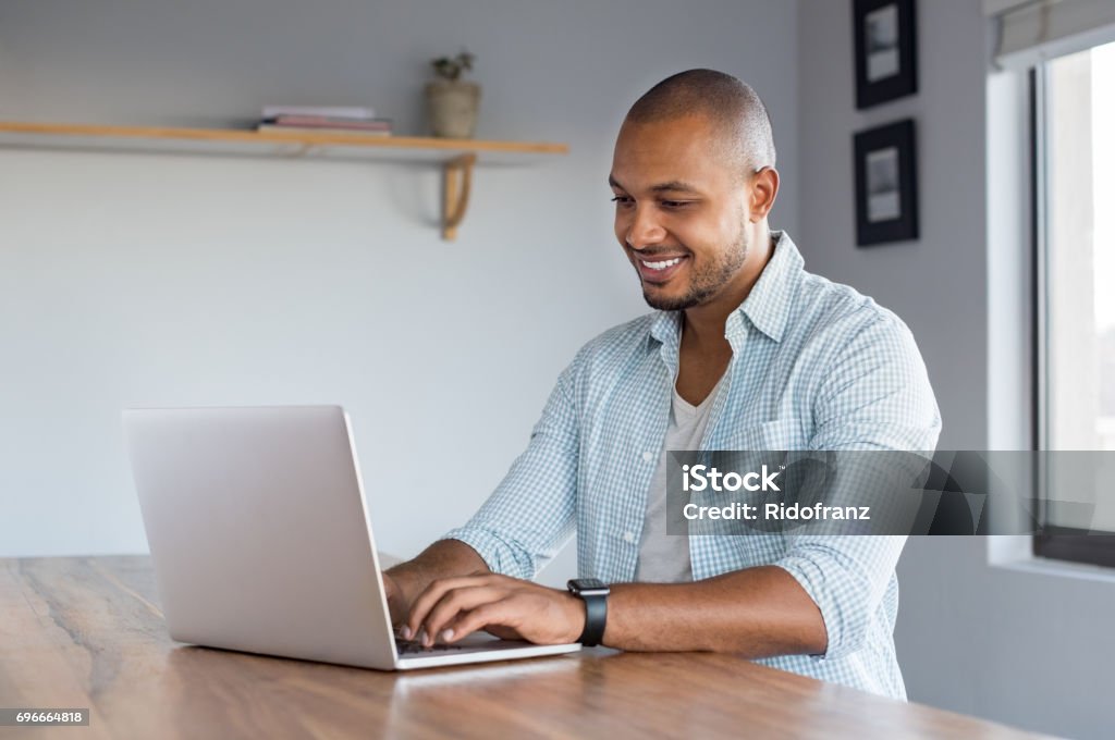 Man working on laptop at home Young casual man working on laptop at home. African businessman typing on computer to send an email. Happy smiling black man surfing the net in a living room. Men Stock Photo