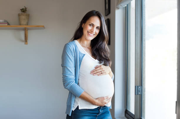 Happy pregnant woman Happy pregnant woman with big tummy relax at home. Portrait of pregnant mature woman standing near the window and caresses her belly. Smiling woman expecting child and looking at camera. pregnant women stock pictures, royalty-free photos & images