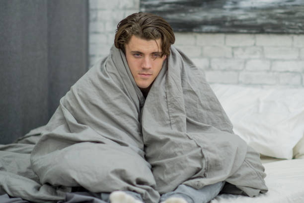 Huddled in Blankets A Caucasian man is sitting in bed. He is sick and getting some rest. He is sitting up and holding a duvet around him, trying to keep warm. cold and flu man stock pictures, royalty-free photos & images
