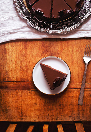 Dark chocolate cake with whipped cream frosting on a silver tray and slice of cake on a plate on a wooden table, top view