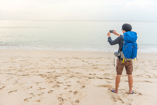 Young Asian backpacker man take photos of beach and sea by smartphone, summer holiday vacation and travel tropical island concepts