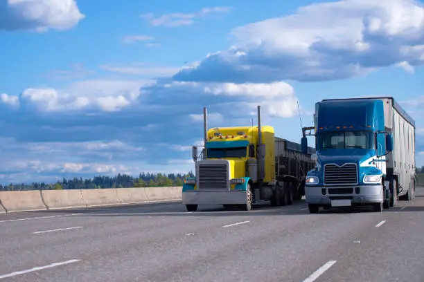 Two semi trucks of various models and manufacturers, a yellow classic American semi truck with a bulk trailer and a blue modern American semi truck with a high trailer for bulk cargo rushing along a wide highway next to each other