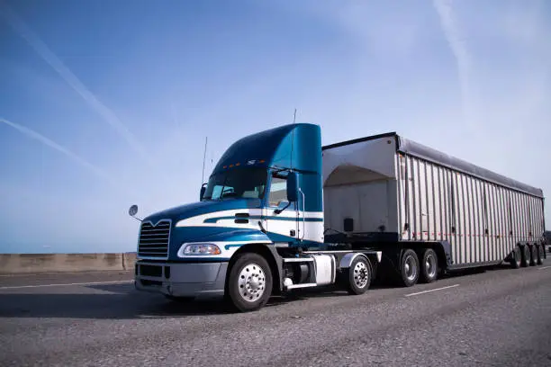 Contemporary big rig blue semi truck with high day cab and covered semi trailer for delivery bulk cargo moving on the wide highway with blue sky and little clouds on background