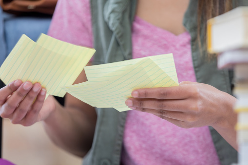 Female college student holds yellow notecards. She is studying for an exam. Focus is on her hands holding the cards.