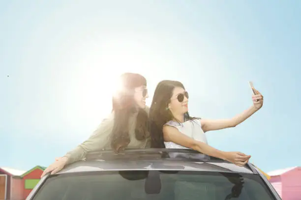 Two young women taking selfie photo with smartphone on a car sunroof near the cottage