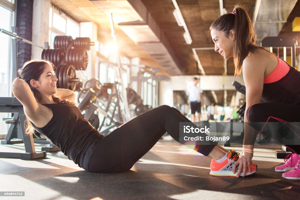 Young woman exercising sit-ups with assistance of female friend in gym. Fitness Instructor Stock Photo