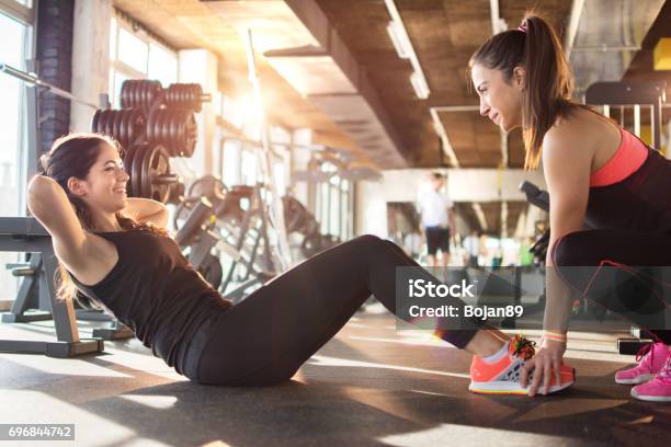 Young Woman Exercising Situps With Assistance Of Female Friend In Gym Stock Photo - Download Image Now