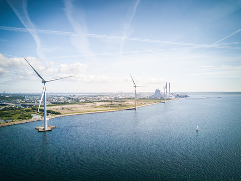 Aerial drone shot of of wind turbines in the ocean at Avedøre, Copenhagen, Denmark. The windmill and sailboat in the foreground stand in contrast to the coal power plant in background. Blue sea and blue sky with warm lens flare.