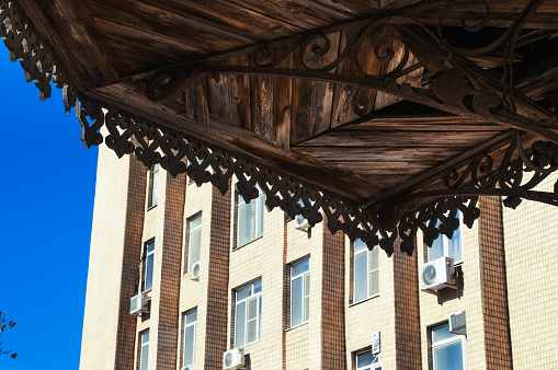 The bottom of the dark balcony of the old Russian house with carved elements of jewelry, against the background of the yellow building of the Soviet era. Astrakhan city, Russia (European part of Russia)