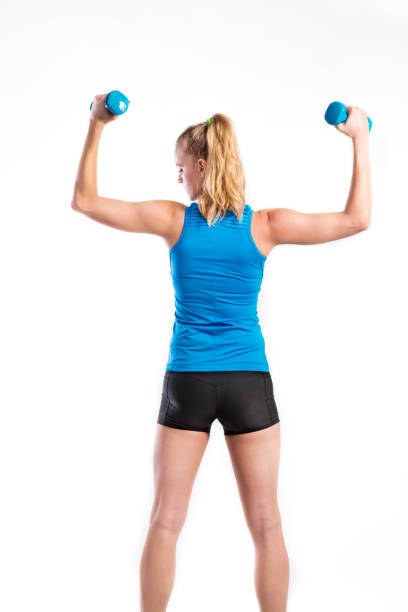 Attractive young fitness woman holding dumbells. Studio shot. Attractive young fitness woman in blue tank top, holding dumbbells. Slim waist, perfect fit female body. Studio shot on gray background. Rear view. blonde female bodybuilders stock pictures, royalty-free photos & images
