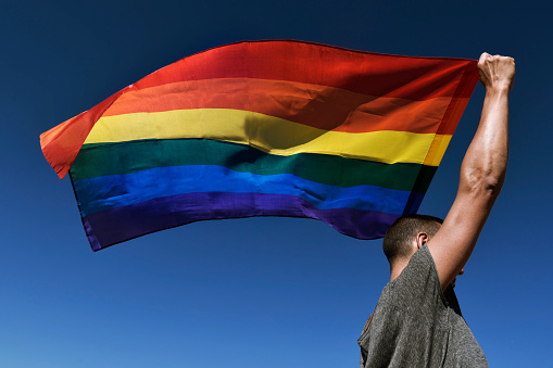 a young caucasian man seen from behind holding a rainbow flag over his head against the blue sky
