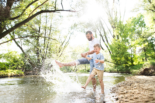 Young father with little boy in the river kicking water, having fun, sunny spring day.