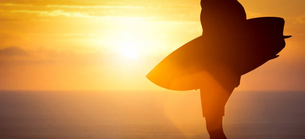 Surfer standing with his surfboard on the beach at sunset over the ocean. Summer time.