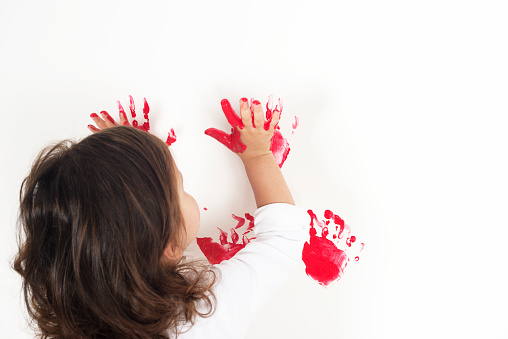 Caucasian girl is painting a white wall with her hands full of red finger painting color.