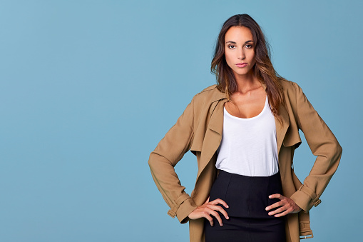 Portrait of beautiful woman standing against blue background. Confident young female is with hands on hip. She is wearing brown trench coat.