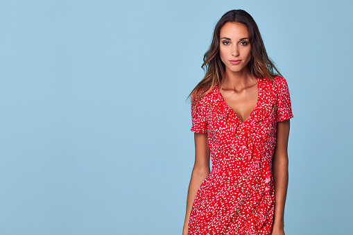Portrait of beautiful female in red sundress. Young woman is standing against blue background. She is with confident look on face.