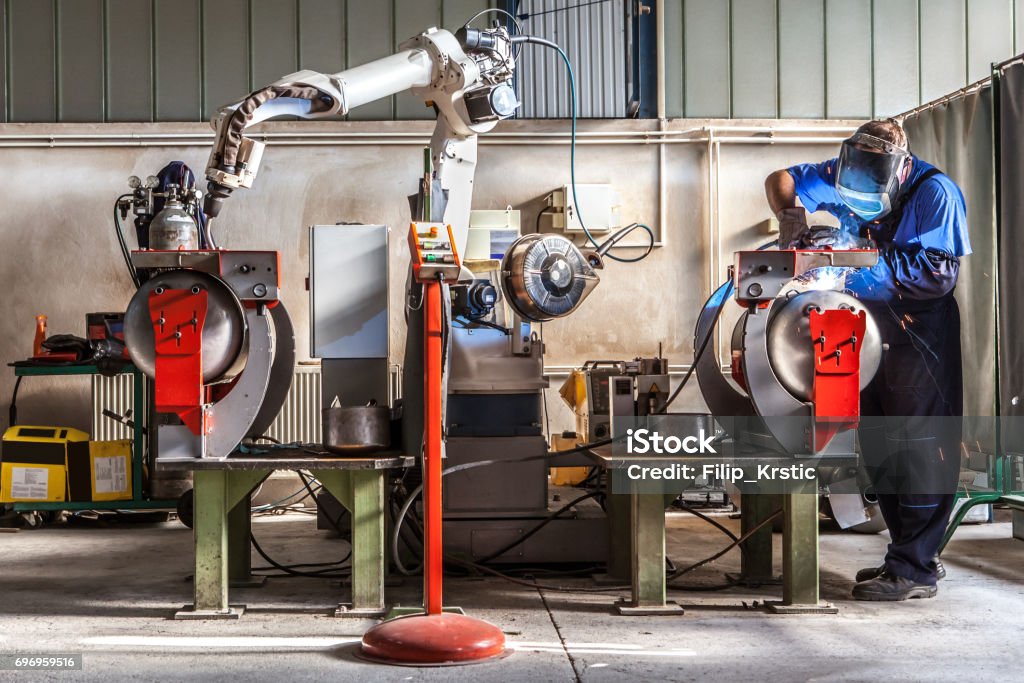 Man and robotic machine work together inside industrial building. The mechanical arm performs welds on metal components assisted by a worker who in turn manages welds manually. Details of the production process. Welding Stock Photo