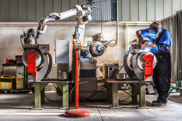 man and robotic machine work together inside industrial building. the mechanical arm performs welds on metal components assisted by a worker who in turn manages welds manually. - manually imagens e fotografias de stock