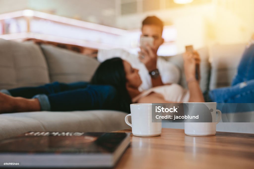 Coffee cups on table with couple relaxing in background Two coffee cups on table with couple relaxing in background on couch. Cups of coffee in front with man and woman at the back at home. Coffee Table Stock Photo