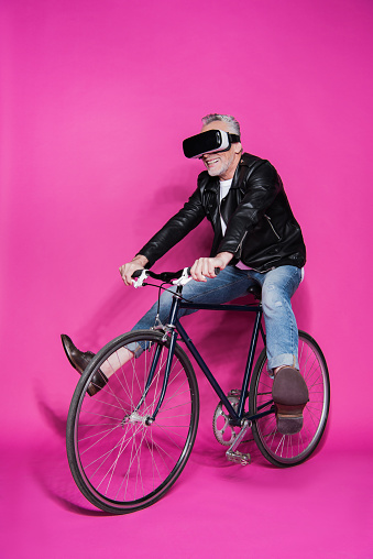 Smiling senior man wearing virtual reality headset and riding bicycle isolated on pink