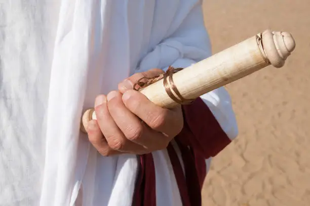 Roman toga, hand of an unrecognizable man holds an ancient or antique book in the form of a vellum or parchment scroll. Sand surface background. Symbol of education, knowledge and science.