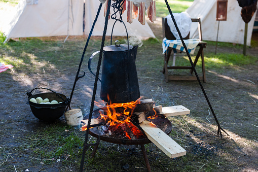 Ancient vintage kettle or a teapot hangs over the open fire in an ancient camp. Slices of bacon hangs over the cattle over the fire, A caldron with peeled off onions stands by the fire, some tents in the background. History reenactment camp.