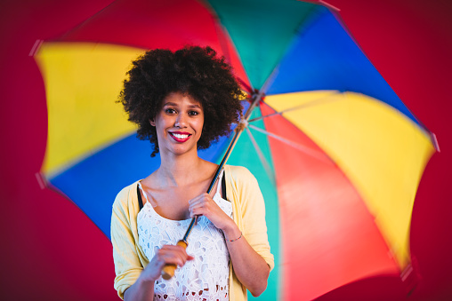 Portrait of young African-American woman looking at camera, holding colourful umbrella and smiling.