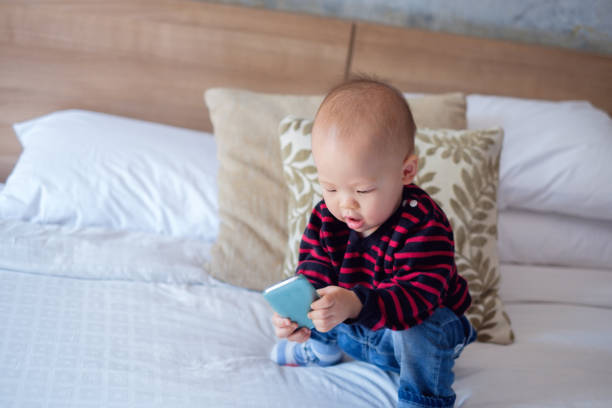 süße asiatische kleinkind baby junge sitzt im bett und hält & smartphone betrachten - phone cradle stock-fotos und bilder