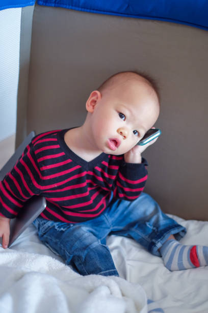 cute asian toddler baby boy sitting in crib and talking on phone & holding tablet - facebook mobile phone telephone smart phone imagens e fotografias de stock