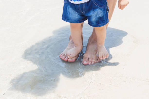 glückliche familie auf einem spaziergang im sommer. niedliche kleine asiatische 1 jahr alt kleinkind baby junge kind mit vater. füße barfuß im sand am strand - child human foot barefoot jeans stock-fotos und bilder