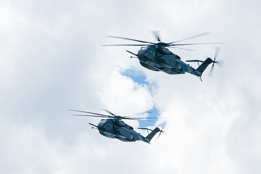 A pair of MH-53 Navy helicopters fly by right to left under broken clouds.