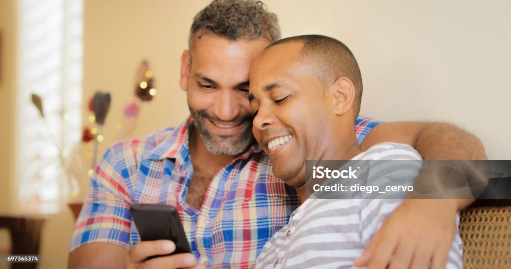 Happy Gay Couple Looking At Pictures On Mobile Phone Homosexual couple, gay hispanic men watching media on mobile phone, sitting on sofa at home. Gay Man Stock Photo