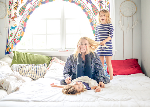 Mother in pajamas with coffee sits on the bed while children jump all around her