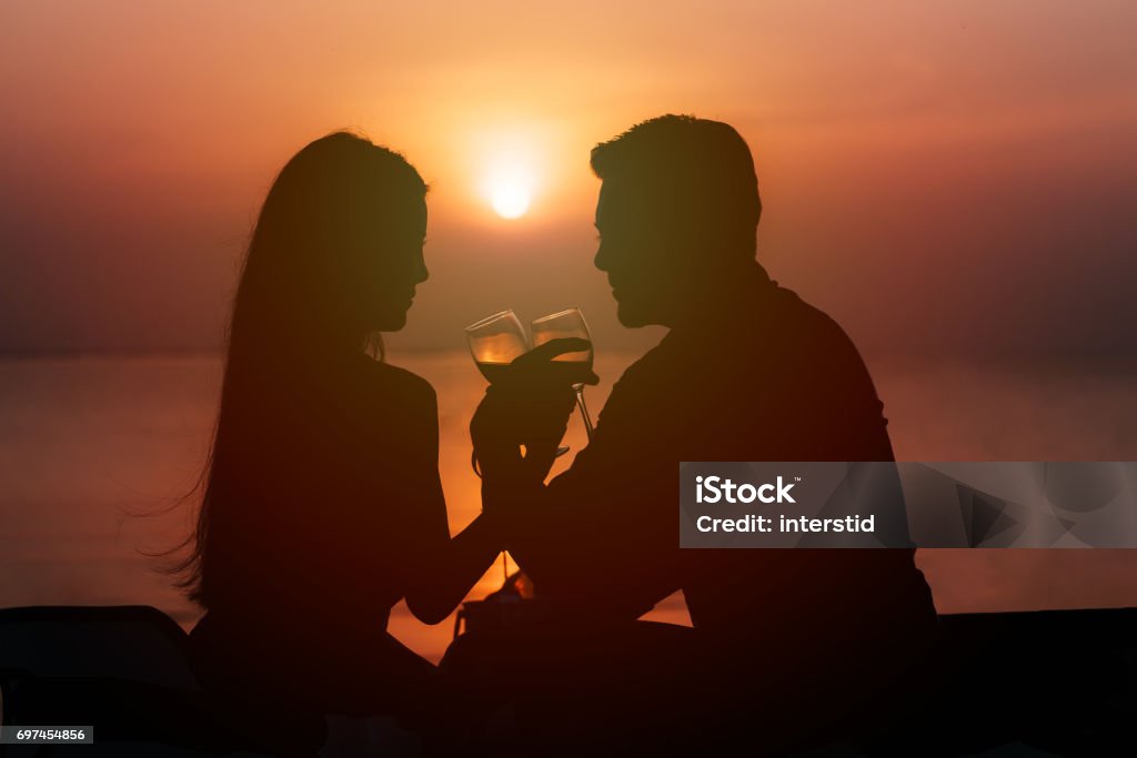 Silhouette of couple in love drinking wine during romantic dinner Silhouette of couple in love drinking wine during romantic dinner at sunset on the beach Couple - Relationship Stock Photo