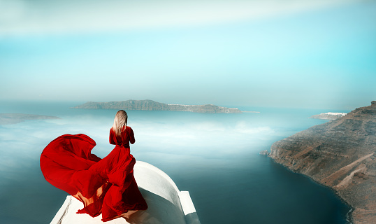 rear view of fashion model enjoying the amazing view over Santorini island. wearing red flying dress.