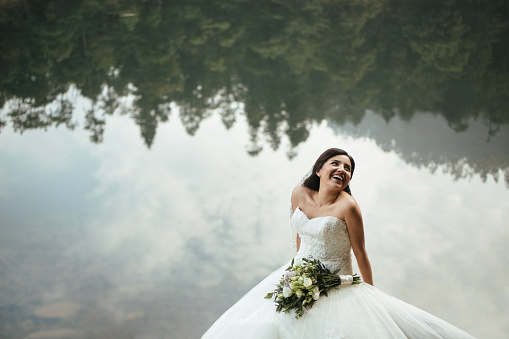 A joyful bride with bouquet looking away and laughing in a horizontal medium shot with lake in background.