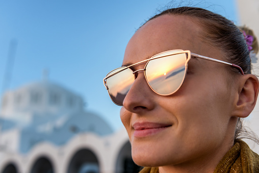 Horizontal color close-up of young woman watching the sunset in Santorini and having the reflection in her mirrored sunglasses. Church in the background.