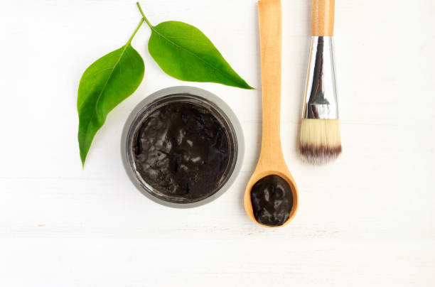 Black clay mineral facial mask, above view mud in jar with spoon and cosmetic brush,  white wooden background. botanical spa treatment stock pictures, royalty-free photos & images