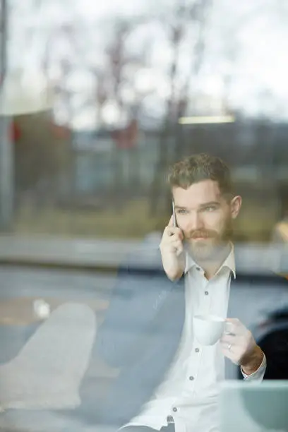 Businessman with cup of tea sitting by window in cafe and calling