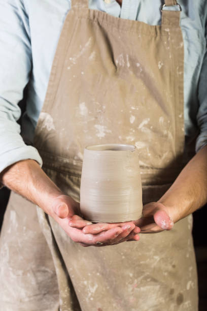 pottery, stoneware, ceramics art concept - young man in a dirty apron stand at workshop, master hands holds cup of unfired clay, male examines product before further processing, front view, vertical stock photo