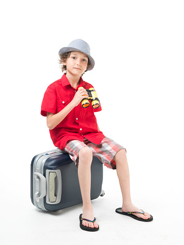 12-13 years old adolescent boy posing with binoculars and suitcase.He is sitting on suitcase and is wearing summer clothes plaid short and red t-shirt.The background is white.Shot in full length in studio with medium format camera.