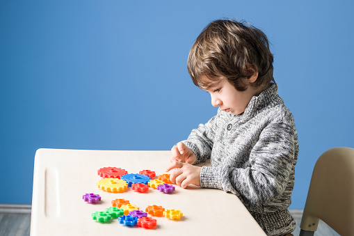 Little boy sitting on school desk and playing with multi colored gears.He has blonde hair.The wall is blue.The model is sitting on the right side of horizontal frame.Shot with medium format camera.