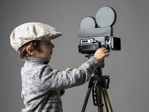 Little boy wearing newsboy cap and gray sweater filming via old fashioned self modified video camera.Cinema film director concept is aimed.The model is smiling.The camera is mounted on tripod.The model is located on left side of frame and background is gray.
