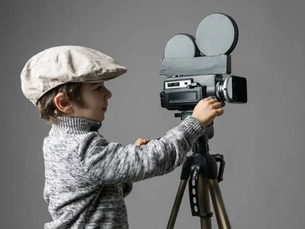 Little boy wearing newsboy cap and gray sweater filming via old fashioned self modified video camera.Cinema film director concept is aimed.The model is smiling.The camera is mounted on tripod.The model is located on left side of frame and background is gray.