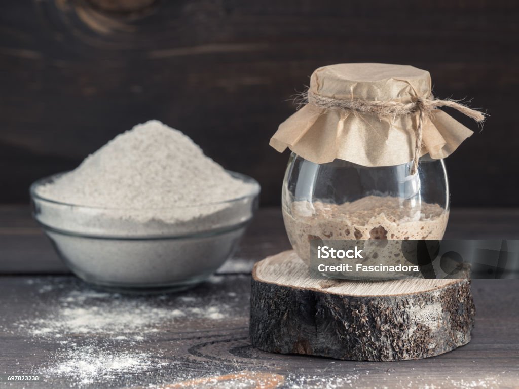 rye sourdough starter and rye flour Active rye sourdough starter in glass jar and rye flour on brown wooden background. Starter for sourdough bread. Toned image. Copy space. Yeast Stock Photo