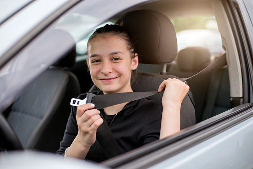 car safety: Happy female teen driver fastening seat belt during the day and looking at camera. transportation and vehicle concept, Hand buckled up with seat belt inside the car