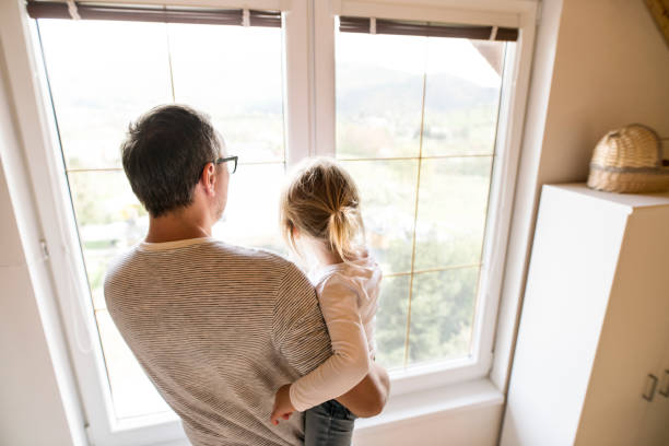 Unrecognizable young father at home standing at the window with his cute little daughter holding her in the arms.